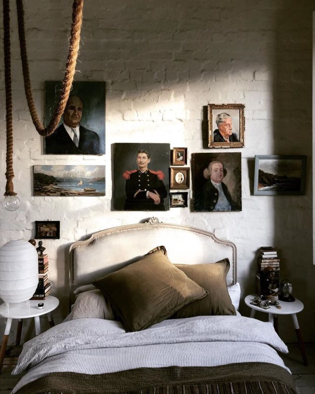 Bedroom with white painted brick wall and nostalgic gallery wall of vintage portraits over bed with brown velvet pillows