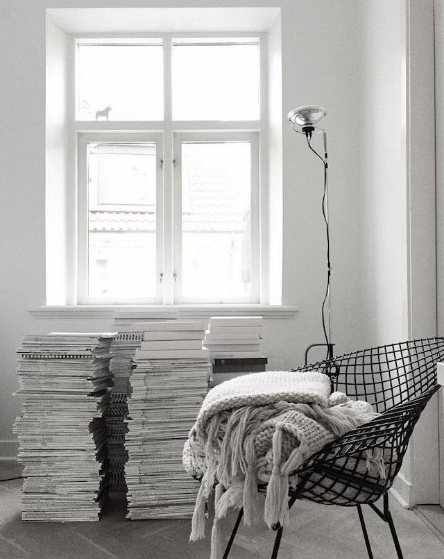 Window with stacks of books on floor and black wire chair piled with cozy blankets