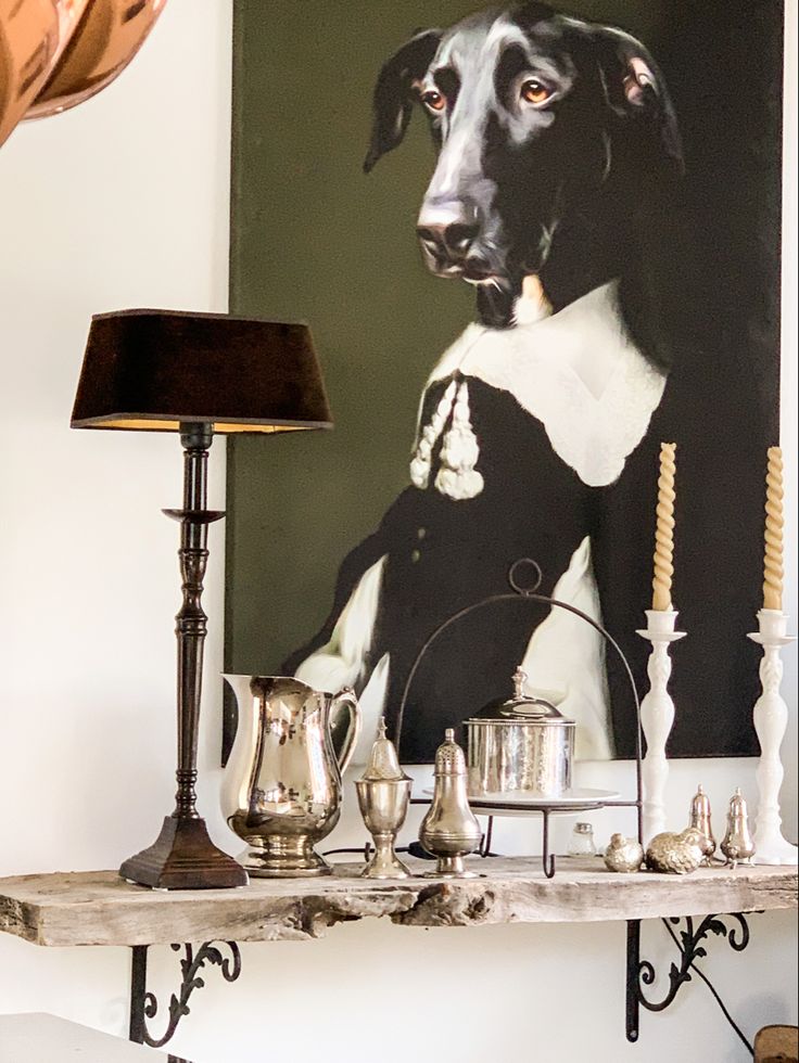 Oversized painting of black and white dog above white wooden ledge with silver and taper candlesticks and black lamp