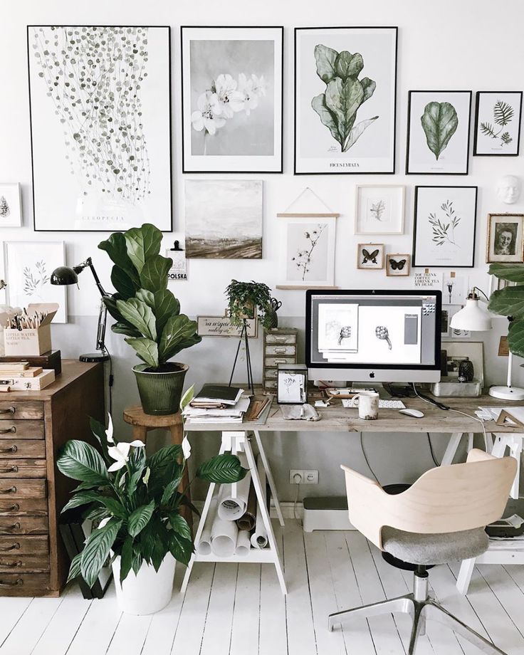 Inviting home office space decorated with botanical art prints on the wall, a large monitor on a white desk surrounded by green houseplants, and a modern wooden chair.