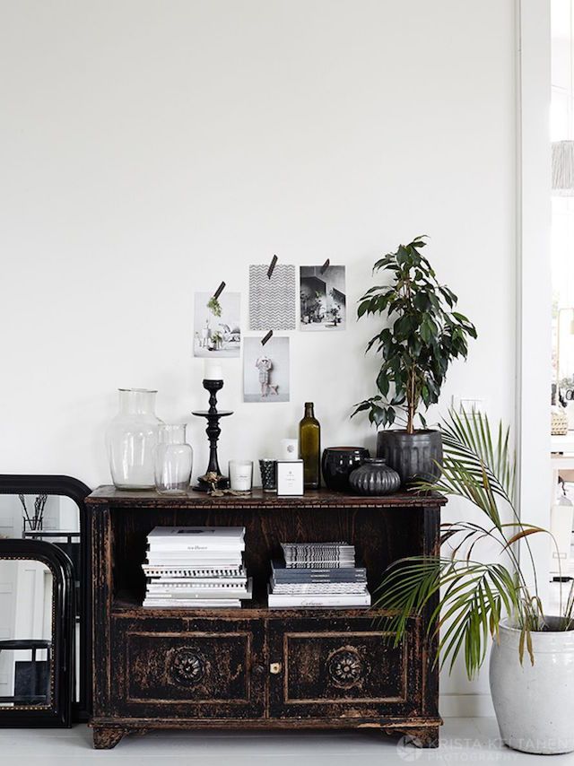 A rustic dark wooden buffet with intricate carvings holds a collection of monochromatic literature, complemented by clear glass pitchers and a solitary green bottle. Above, a curated selection of grayscale images creates a simple wall gallery beside a thriving houseplant.