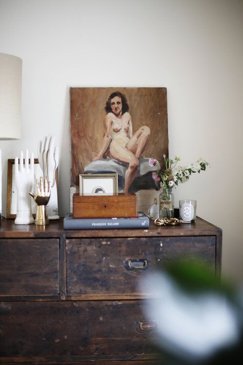 An antique wooden dresser displays a curated collection of items including a vintage oil painting of a seated woman, a stack of books with 'PARADISE ISLAND' on the spine, a white ceramic hand sculpture, a lamp with a white shade, and small decorative objects including a clock and a vase with fresh flowers.