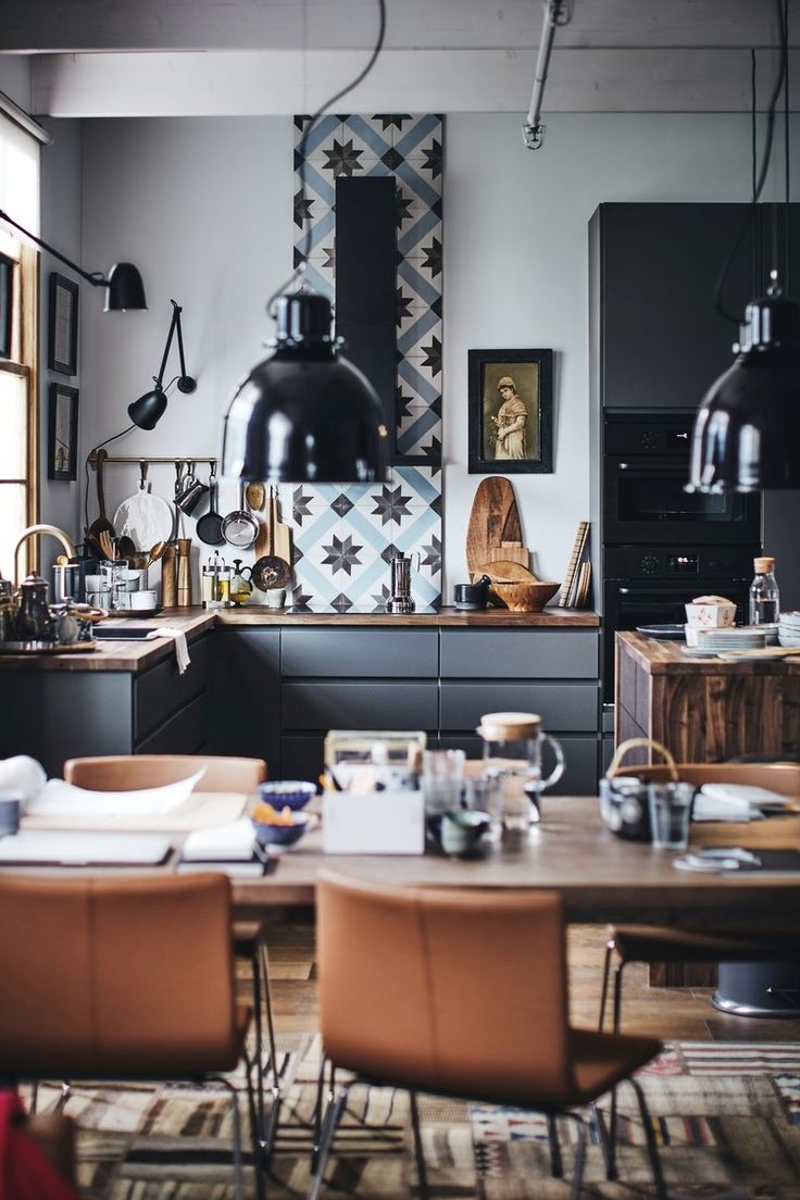 A stylish dining area with an industrial chic aesthetic, featuring a wooden table with tan leather chairs on a patterned area rug. Overhead, large black pendant lights hang, casting a warm glow. The kitchen in the background boasts sleek dark cabinetry, enhanced by a striking tiled backsplash with geometric patterns. Vintage culinary tools and modern appliances share the space, reflecting a blend of traditional charm and contemporary design.