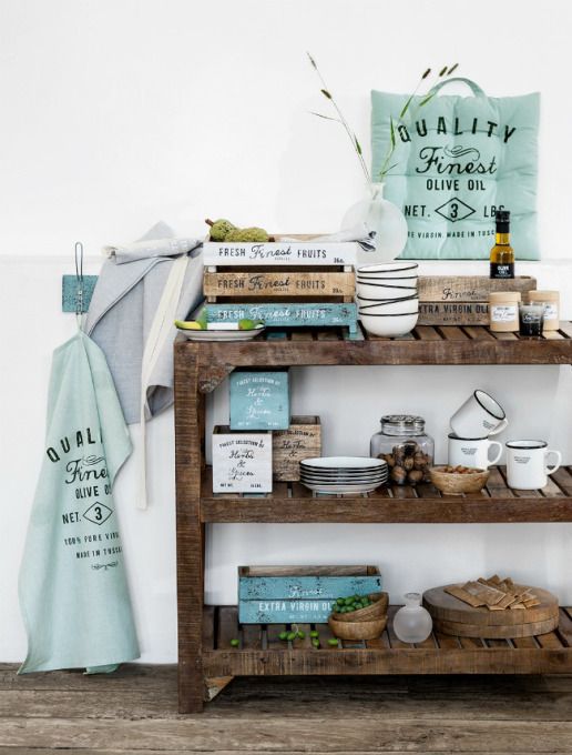 A rustic wooden open shelf kitchen organizer displaying various kitchenware, including bowls, plates, and jars, with decorative items such as a cushion with 'Quality Finest Olive Oil' print, and a 'Dual Fine' labeled towel hanging on the side.