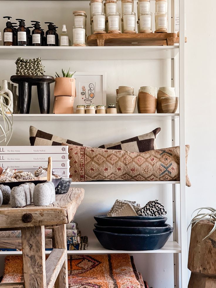 An inviting retail shelving display showcasing a variety of home goods including black pottery, neatly arranged skincare products, and artisanal ceramics, with rustic wooden elements and a cozy, earth-toned cushion for a warm, autumnal feel.