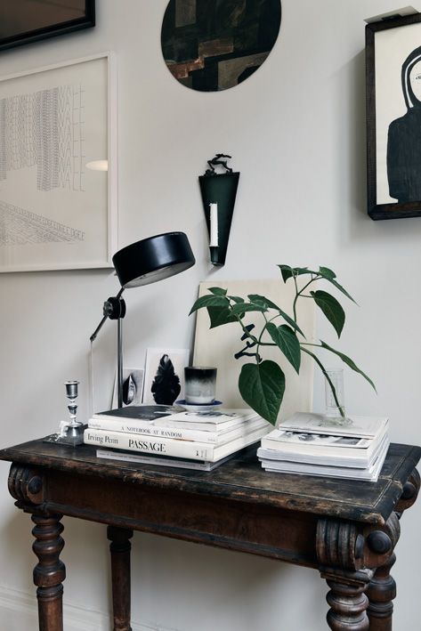 A tastefully decorated vignette featuring a rustic wooden console table adorned with a carefully curated selection of items. A black adjustable desk lamp provides a modern contrast to the antique table. Stacked books, including titles such as 'A Journey of Wonders' and 'PASSAGE', create a base for a green potted plant, which adds a touch of life and color to the composition. The wall above is decorated with framed art pieces, including a text-based graphic and two abstract paintings, contributing to the room's artistic and eclectic charm.