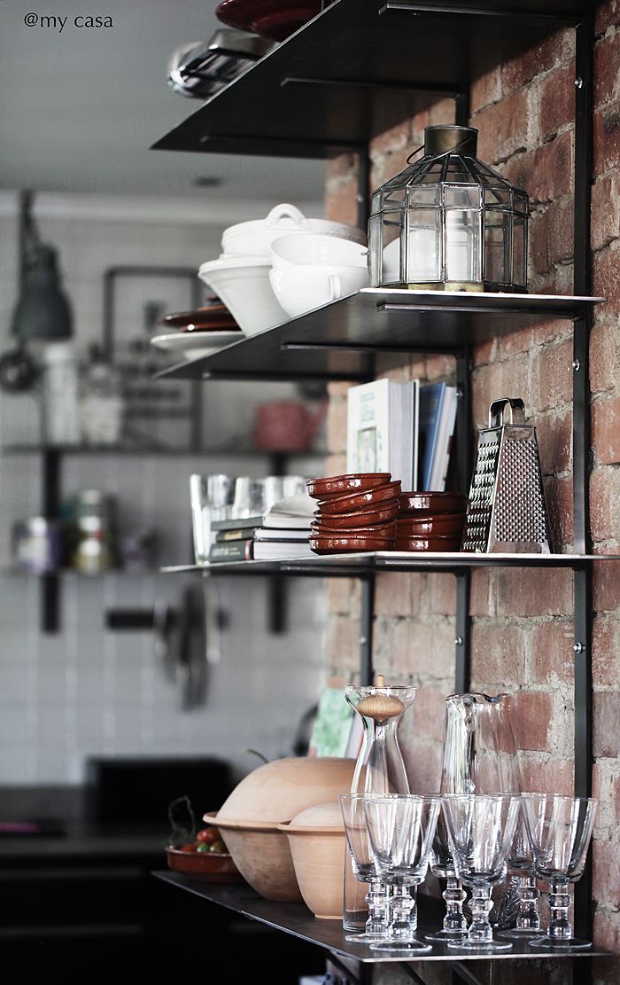 Close-up of open kitchen shelving against a rustic brick wall. The shelves hold an array of kitchen items including white serving bowls, a grater, a stack of brown ceramic plates, cookbooks, and clear glassware featuring wine glasses and a decanter. The arrangement is practical yet aesthetically pleasing, highlighting a balance between functionality and homey decor.