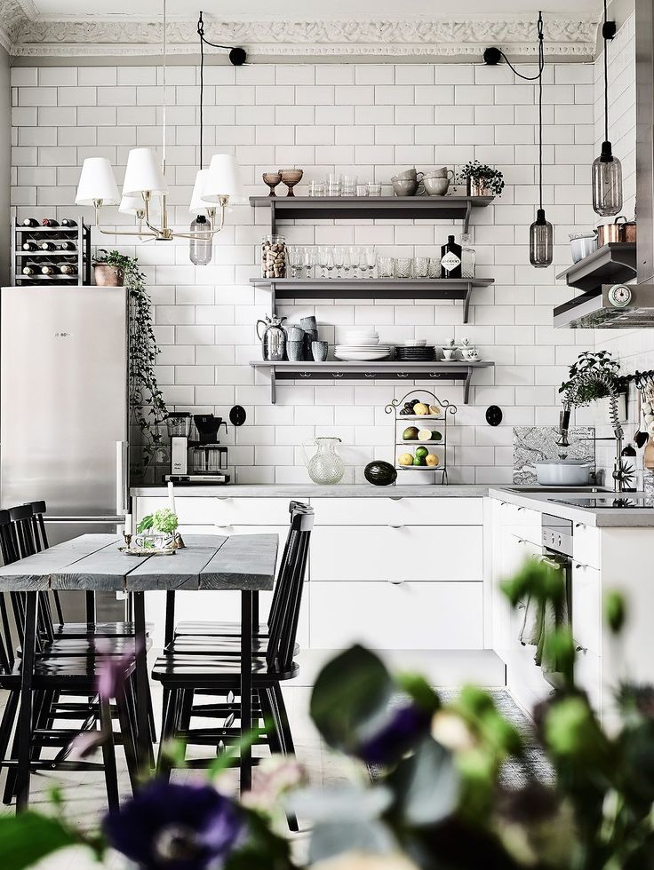 A sleek and modern kitchen with white subway tile walls and open shelving displaying a variety of dishes, glasses, and kitchenware. A black dining table with matching chairs sits in the foreground, adorned with a small plant centerpiece. Elegant pendant lights hang above, and a stainless steel refrigerator adds to the contemporary look. The space is accented with subtle touches of greenery, bringing a sense of life and freshness to the chic, monochromatic color scheme.