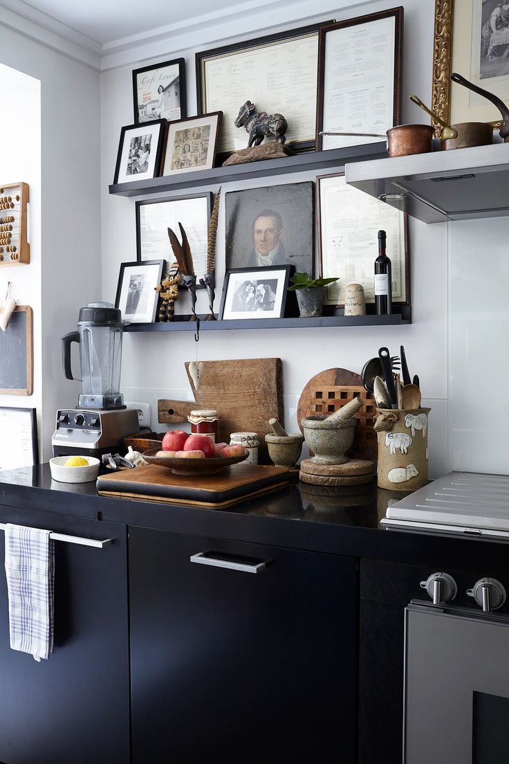 Kitchen counter and shelves with a variety of items including framed pictures, a blender, cutting boards, apples on a wooden stand, and kitchen utensils in a holder.