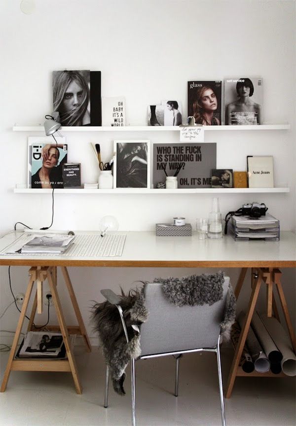 A minimalistic home office setup featuring a wooden desk with A-frame legs and a grey upholstered chair with a fluffy grey seat cover. Above the desk, two white shelves display a curated selection of monochromatic photographs and magazine covers with bold typography. On the desk, there is a white keyboard, a pair of glasses, a stack of books, and various office supplies, creating a clean and stylish workspace.