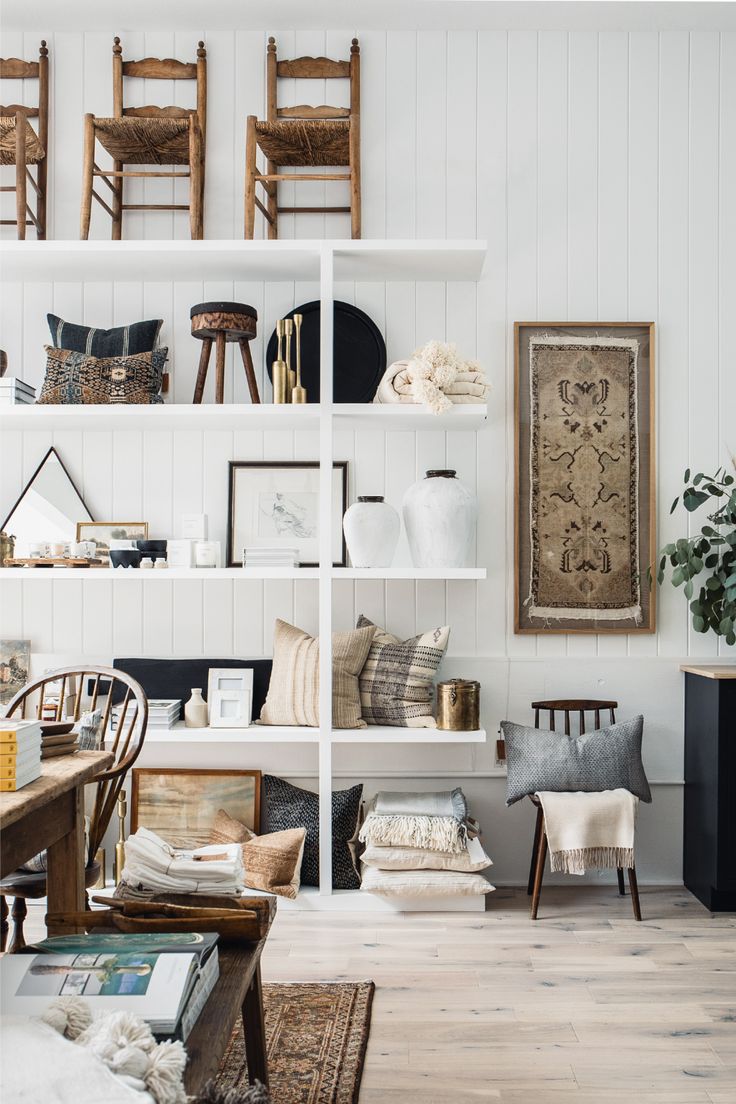 A cozy and stylish living space featuring a white shelving unit adorned with decorative items including vintage chairs, framed art, vases, and assorted pillows, with a traditional rug on the wooden floor.