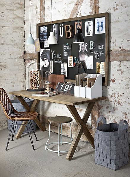 An eclectic home office corner with a rustic vibe, showcasing a wooden trestle desk and a woven rattan chair. A large inspiration board filled with assorted monochrome images, quotes, and pendant lights hangs on a textured brick wall. A wooden stool, decorative pottery, storage boxes, and a woven grey felt basket complement the creative and vintage aesthetic of the space.