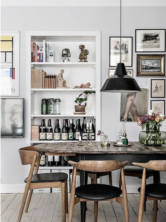 A cozy dining area with a rustic wooden table and four mid-century modern chairs. A black pendant light hangs above, and a shelving unit is filled with books, green glassware, and decorative items. A selection of bottled beverages is displayed on the table alongside a green lamp and a vase with fresh flowers, creating an inviting and personal space.