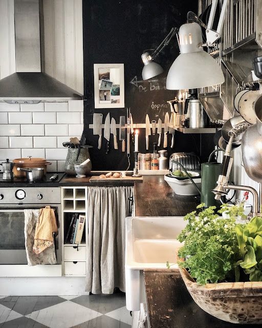 A cozy and rustic kitchen scene with white subway tiles and a chalkboard wall featuring handwritten notes. A large farmhouse sink is nestled beneath a window, with a wooden basket of leafy greens at its side. The kitchen boasts a mix of modern and vintage elements, including a stainless steel stove with a copper pot, a green stand mixer, and various hanging utensils. A unique wooden chandelier with candles adds warmth to the space.