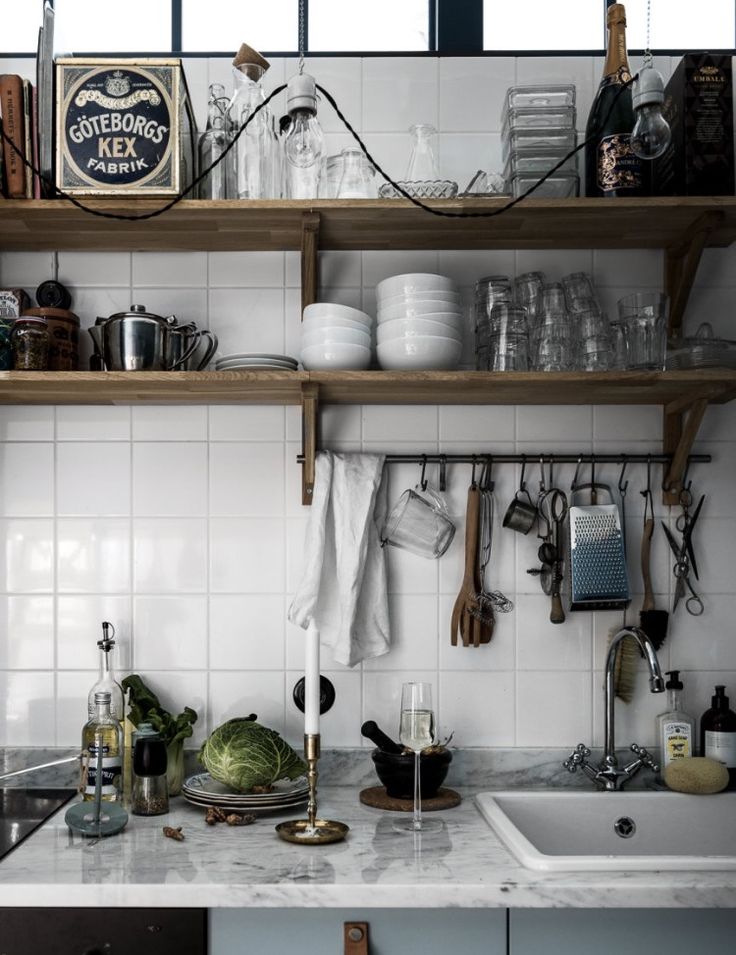 A stylish kitchen counter with open wooden shelves above, displaying a mix of white dishes, glassware, and a vintage Göteborgs Kex biscuit tin. Hanging utensils, a bottle of olive oil, a candlestick, and fresh green cabbage add a homely touch. The monochromatic color palette is accented with natural wood tones and hints of greenery, creating a functional yet inviting culinary space.