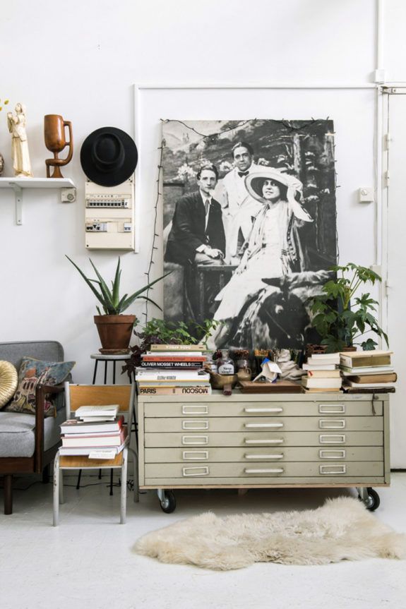 A cozy, vintage-inspired corner featuring a large black and white photograph of three people taped to the wall, an assortment of potted plants, a mid-century gray sofa with decorative pillows, and a metal flat-file cabinet stacked with books and eclectic decor. A sheepskin rug lies on the floor, and various unique items like a wooden wall sculpture and a black hat add personality to the space.
