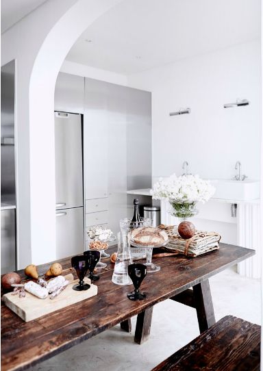 An elegantly simple kitchen interior with a rustic wooden table set with bread, cheese, wine glasses, and a bottle, complemented by a bouquet of white flowers in the background and modern kitchen appliances.