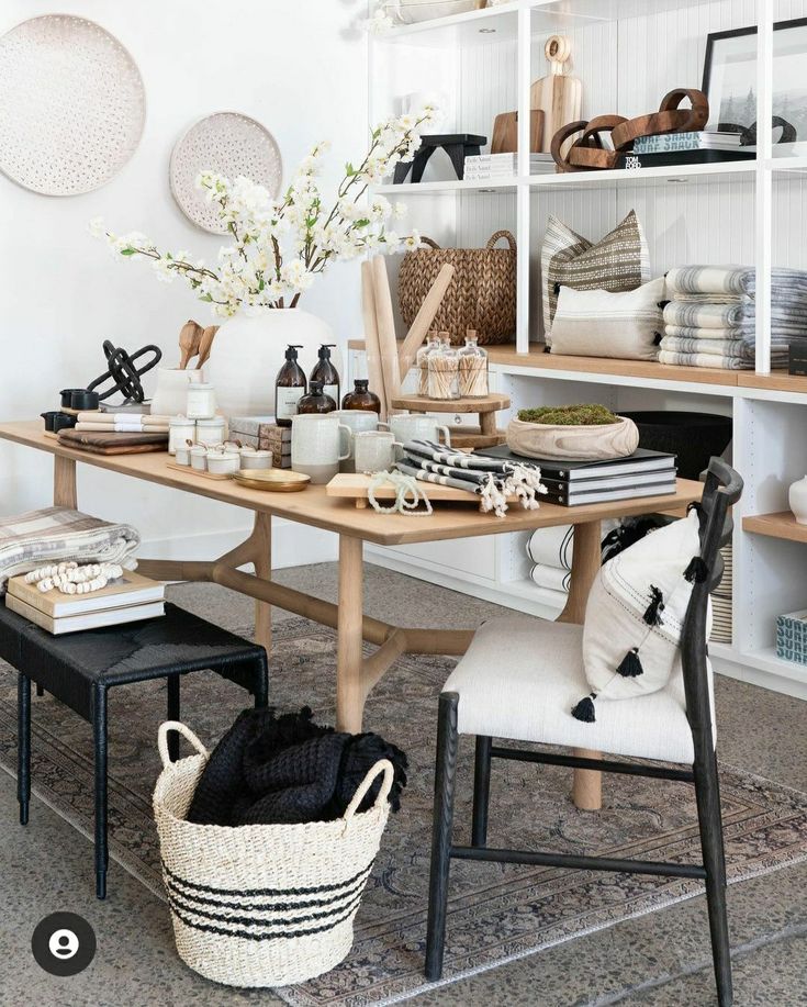 A well-organized home office space with a natural wood desk, surrounded by white chairs with black accents, and a woven basket on a patterned rug. The room is decorated with wall-mounted ceramic plates, a vase of blossoming branches, and shelves showcasing an array of home goods and textiles.