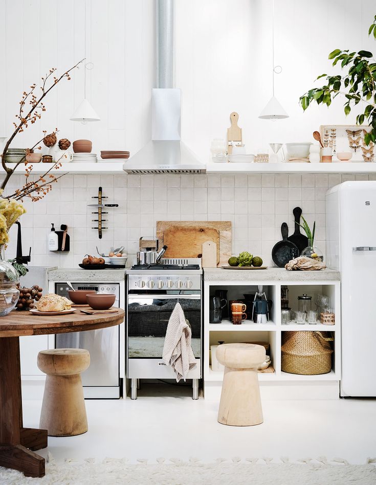 A bright and inviting kitchen space with white wooden panel walls and a fluffy white rug. The area is styled with an earthy palette, featuring a round wooden dining table with matching stools. Open shelving displays a curated collection of pottery and glassware, while a white modern range hood hangs above a stainless steel stove. Natural elements like a branch with blooming buds and green houseplants add a touch of organic warmth to the scene.
