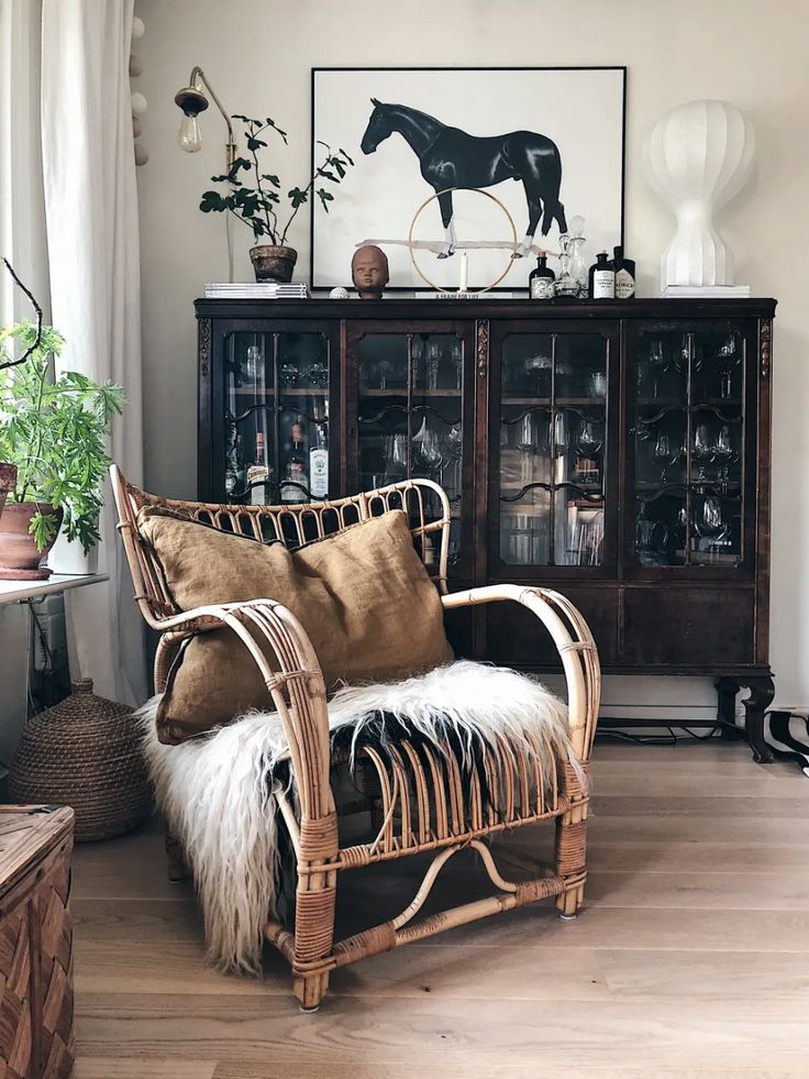 A cozy nook featuring a rattan armchair adorned with plush cushions and a sheepskin throw, beside an antique wooden cabinet displaying various items, all under a framed horse illustration.