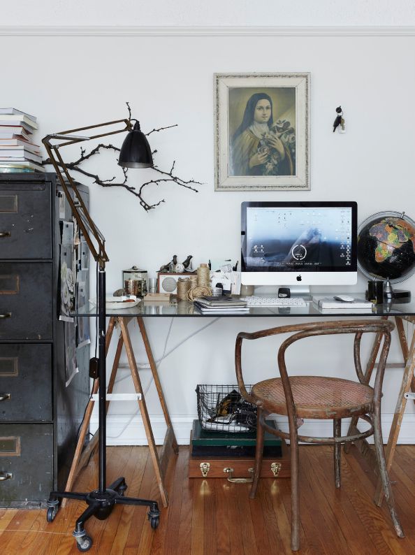 A cozy and artistic home office corner featuring a wooden desk with a rustic finish and a bentwood chair. On the desk sits an iMac with a nature-themed wallpaper, flanked by miscellaneous items like a mug, notebooks, a small globe, and stationary. Above the desk hangs a framed portrait resembling the Mona Lisa. To the left, a black anglepoise lamp extends over the workspace, and a tall, multi-drawer metal filing cabinet adds an industrial touch. The room is bright with white walls, and a branch decor piece adds a natural element to the space.