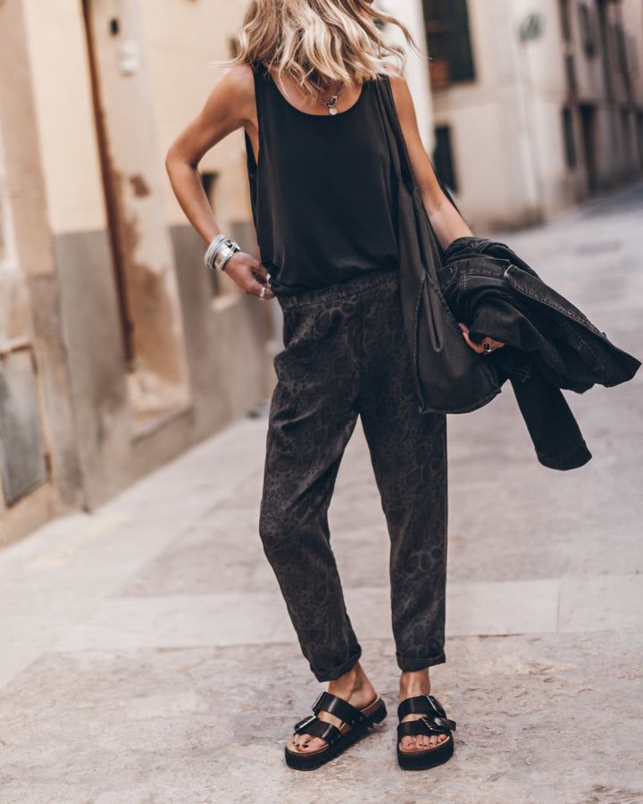 A person in a casual chic outfit with a black top, patterned trousers, and sandals, holding a jacket and walking on a city street.