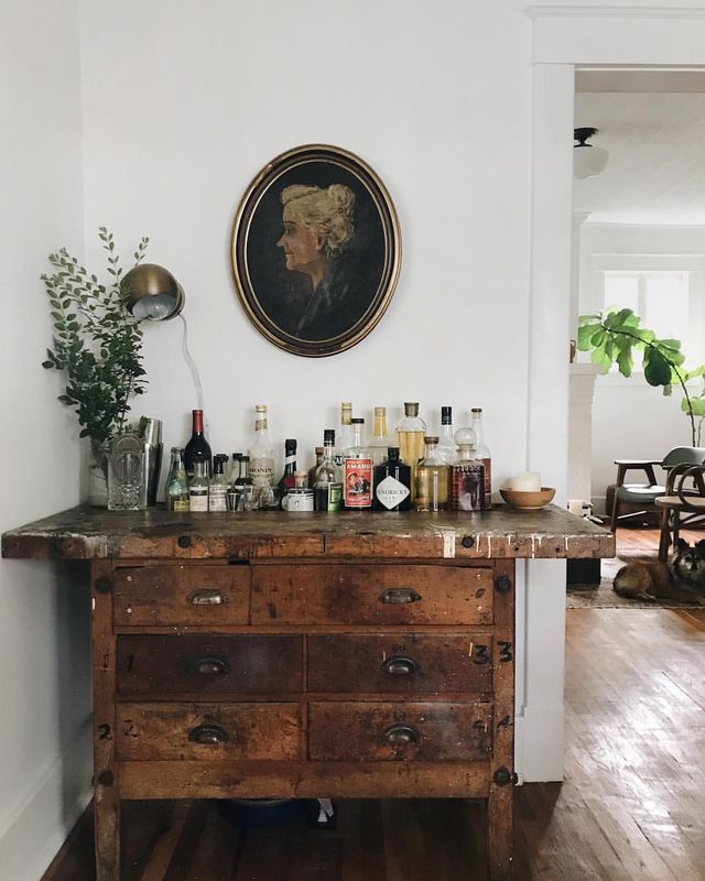 Vintage-inspired home bar setting featuring a rustic wooden apothecary chest adorned with a collection of assorted liquor bottles. Above it hangs a classical portrait in an oval frame, adding a touch of historical elegance to the space.
