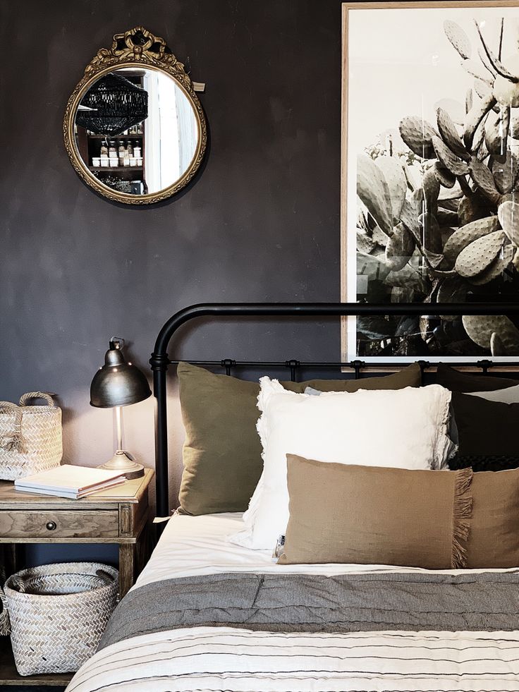 A cozy bedroom corner with a metal bed frame, layered pillows, a bedside lamp, a wicker basket, and a round mirror on a dark wall.