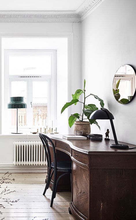 A serene and minimalist workspace set against a backdrop of crisp white walls with elegant crown molding. The room features a classic wooden roll-top desk with a deep patina, accompanied by a simple black chair. On the desk, a potted fiddle-leaf fig plant adds a touch of green, and a vintage-style black desk lamp provides task lighting. A round, frameless mirror on the wall reflects a glimpse of the room, adding depth. The large window with white trim floods the space with natural light, highlighting the warm wood floors. Just out of the window's view, a green pendant lamp casts a cozy glow.