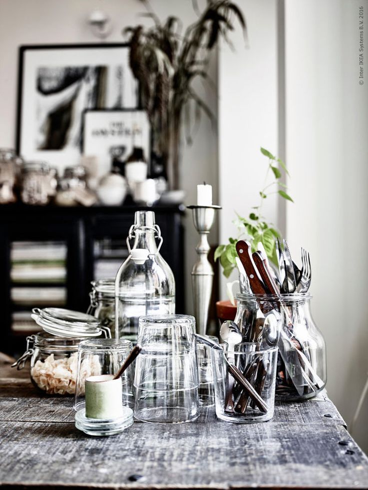 A contemporary kitchen corner featuring a clean and minimalist design. A modern kitchen extractor hovers over a countertop adorned with wooden cutting boards, a potted herb, and cooking utensils. The foreground shows a part of a dining table with a small potted plant, a black candlestick, and a clear jar with budding branches, evoking a serene and inviting atmosphere.