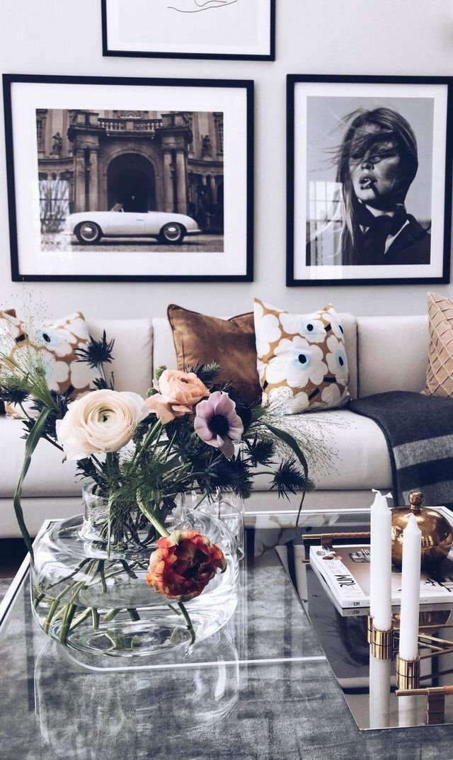 A well-decorated living room featuring a floral arrangement on a coffee table, with art and photographs on the wall, and elegant throw pillows on a couch.