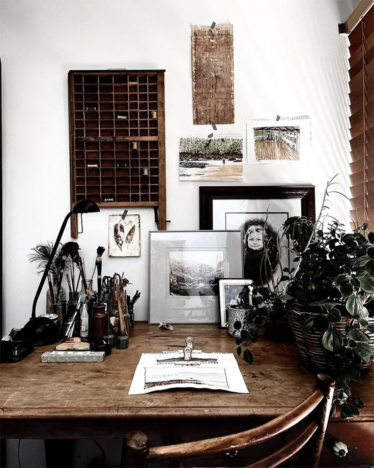 An artist's workspace with a wooden table, various art supplies, framed artwork, and a potted plant, against a wall decorated with assorted pictures.
