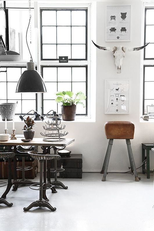 An industrial-chic workspace illuminated by natural light from large windows with black frames. A glossy concrete floor reflects a vintage-style metal desk with unique cast iron legs, surrounded by an eclectic mix of objects including a large black pendant lamp, a rustic brown leather stool, and a skeletal bull's head mounted above framed monochrome art prints.