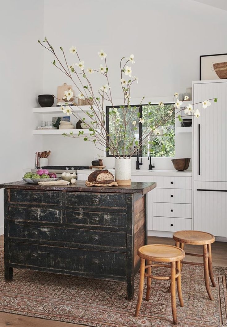A rustic kitchen interior featuring a distressed black island, two wooden stools, white cabinetry, a persian rug, and a large vase with branches and white blossoms on the countertop.