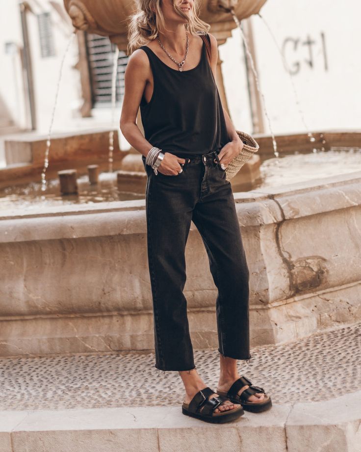 Woman in black sleeveless top and cropped jeans standing by a fountain.
