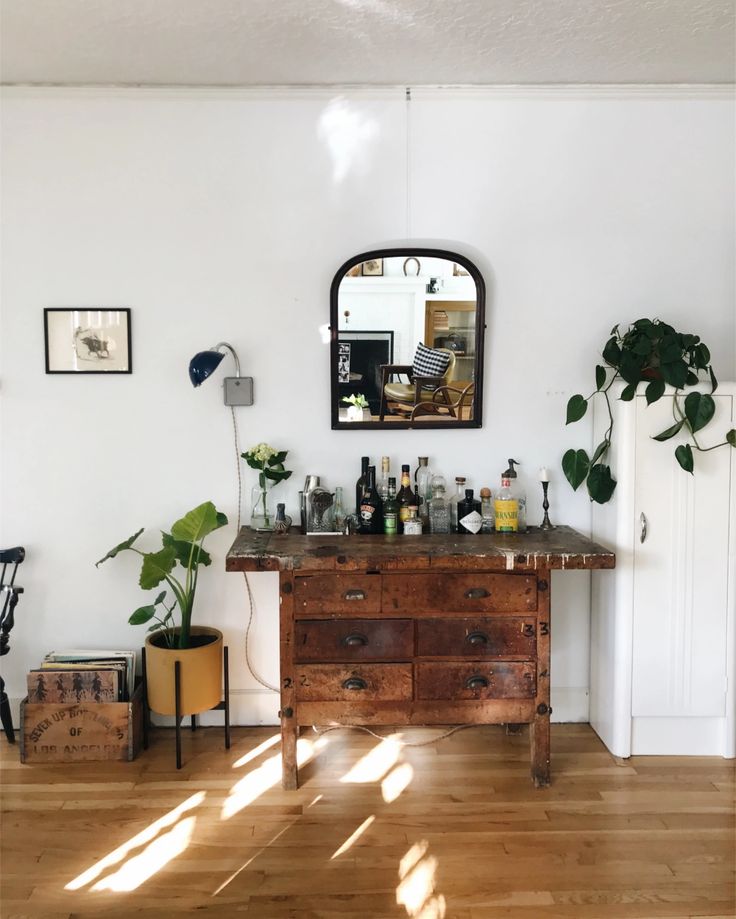 A vintage wooden dresser with various bottles on top, positioned against a white wall with a mirror hanging above it, reflecting an interior space. a potted plant sits to the right, sunlight casts shadows on.
