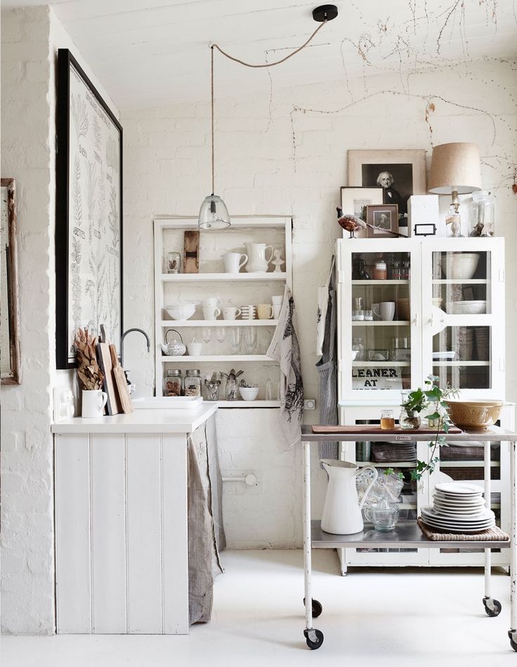 A bright and rustic kitchen corner with white brick walls, featuring a glass cabinet, shelves with various items, and a mobile island cart.