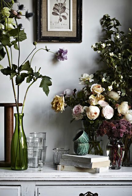 A vintage-style interior with an assortment of flowers in various vases, books, and glassware on a distressed white wooden table.