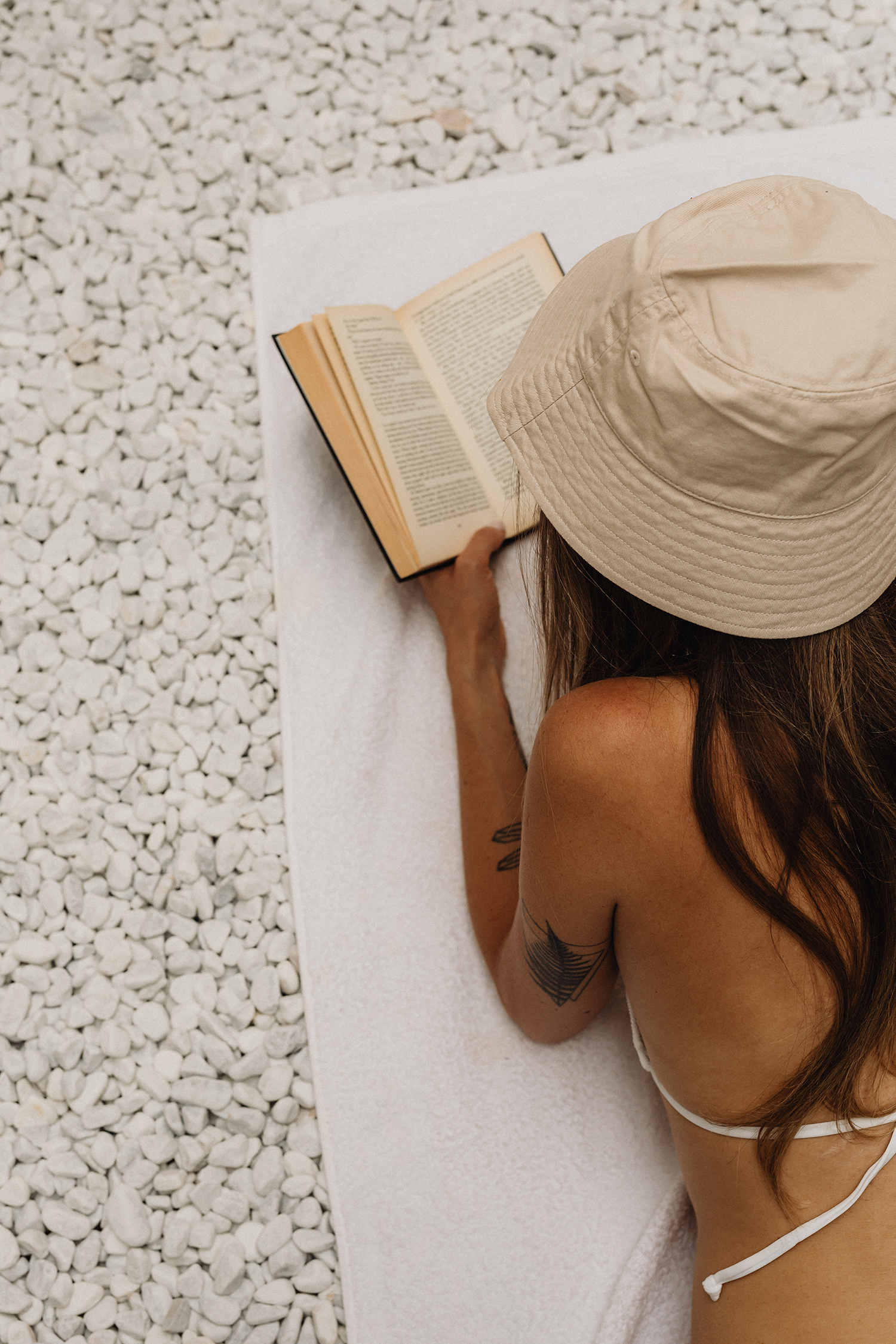 Brunette woman lying on stomach in sand with straw hat reading book
