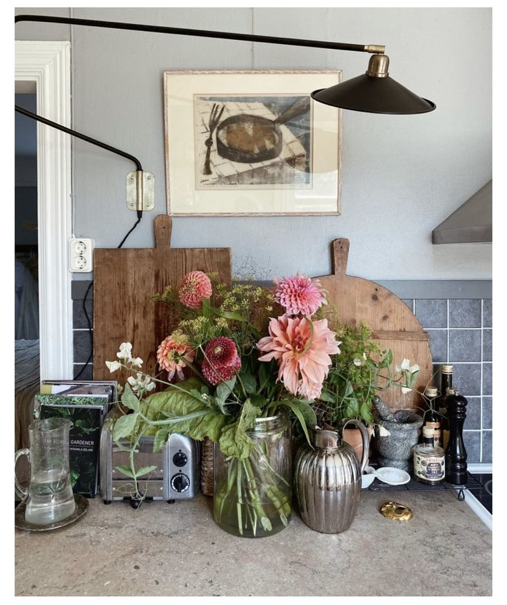 A cozy kitchen corner with an assortment of flowers in vases, wooden cutting boards, and vintage decor items under a wall-mounted picture and lamp.