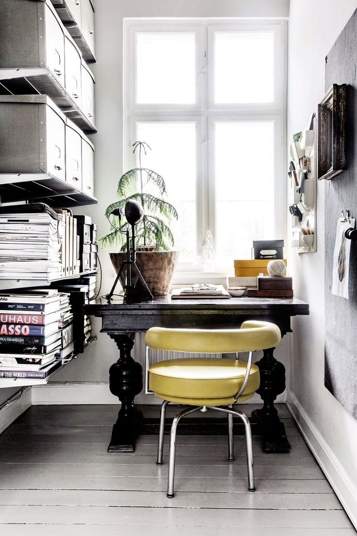 An eclectic study space with a vintage vibe, featuring a heavy, ornate black desk paired with a mustard-yellow mid-century modern chair. The desk is adorned with neatly stacked books, a classic desk lamp, and an assortment of trinkets. A wall-mounted shelf to the right holds additional knick-knacks and office supplies. A tall metal filing cabinet with multiple drawers occupies the space to the left, towering over a pile of large art books on the floor. A lush fern adds a splash of greenery beside the bright, natural light coming in from the tall, white-framed windows, contrasting with the soft grey walls and light grey wooden flooring.