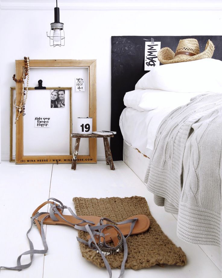 A cozy bedroom corner featuring a white bed with a knit throw, a straw hat resting on the bed, a small wooden stool, a floor lamp, and wall art, alongside a pair of sandals and.