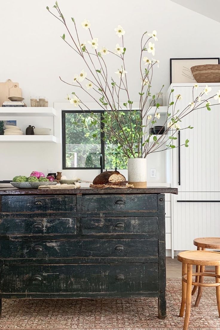 A cozy and rustic corner of a home featuring a distressed black wooden dresser, topped with a ceramic vase of delicate white dogwood branches. A loaf of artisan bread and fresh green artichokes rest casually on the counter, contributing to the room's farmhouse charm. Natural light filters in through the window, illuminating the space and the open shelving with neatly arranged dishware in the background.