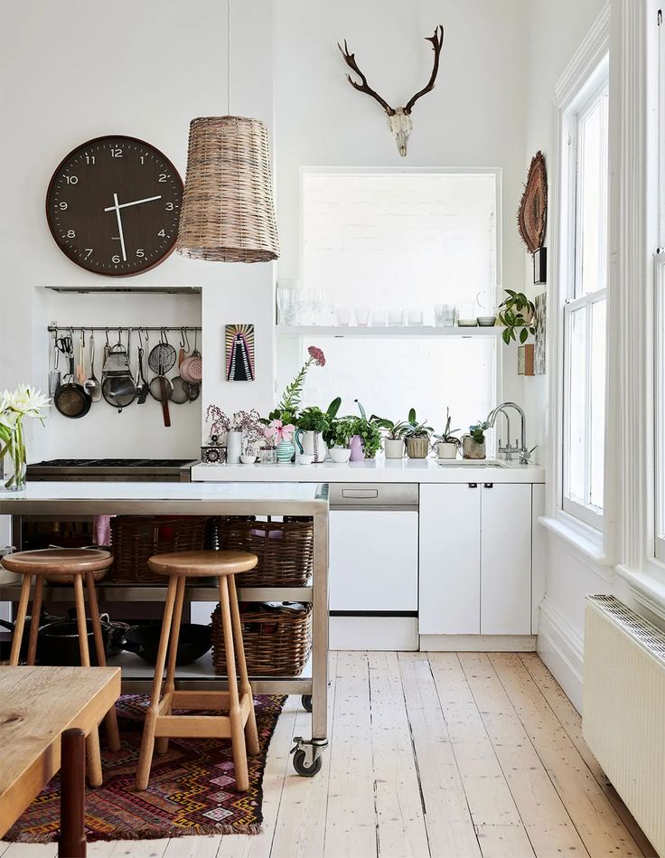 A bright and eclectic kitchen with white walls and painted wooden floors. The space features a large brown clock, a wicker pendant light, and a mounted deer head adding a rustic touch. Below, a functional area includes pots and utensils hanging against a backsplash, beside a countertop hosting an array of potted plants and silver kitchen faucets. Two wooden bar stools tuck under a modern island with storage baskets, and a colorful woven rug adds warmth to the room.