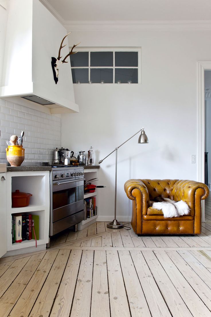 Cozy corner in a Scandinavian-style kitchen featuring a plush, tufted leather armchair with a white fur throw, a classic stainless steel stove nestled next to white subway-tiled walls, and a vintage-style metal floor lamp. Above, a rustic antler mount adds character to the space, while a small transom window allows for additional light. The room is completed by a warm, rustic wood plank flooring.