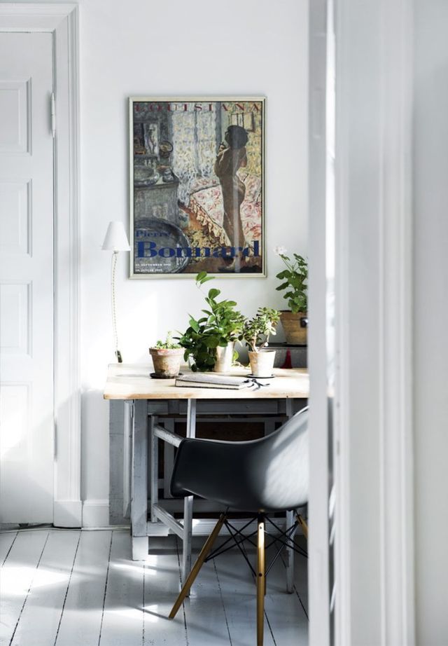 A small, tranquil workspace nestled in a bright room with white walls and painted floorboards, highlighted by the sunlight beaming through a window. A minimalist black chair with golden legs is tucked under a rustic wooden desk, which is home to a collection of potted plants, contributing to a bohemian vibe. A large framed poster of Pierre Bonnard's art adds a touch of cultural sophistication to the space.