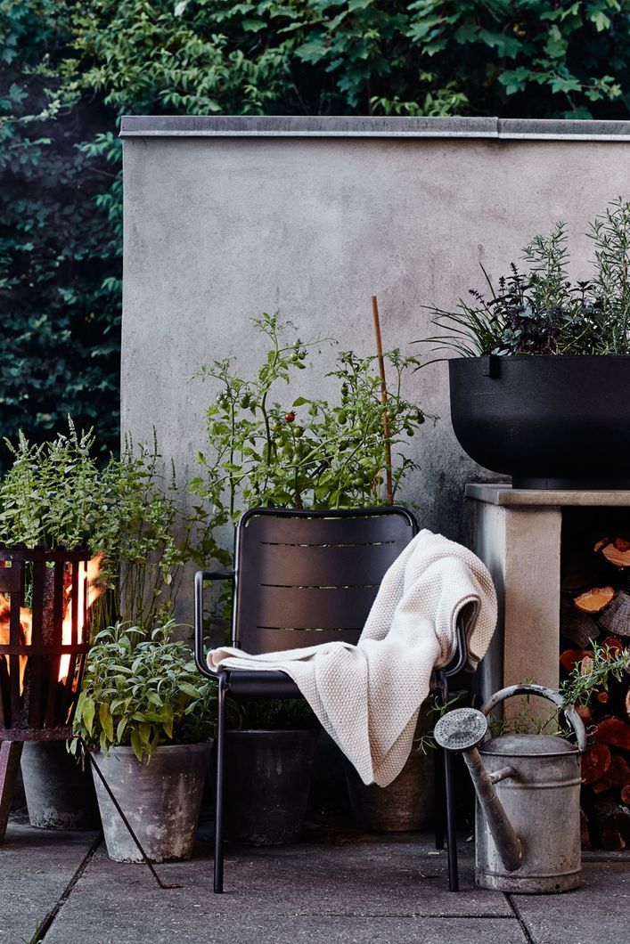A cozy outdoor seating area at dusk with a modern metal chair draped with a cream textured throw blanket. To the left, a rustic fire basket glows warmly next to potted greenery, including a tomato plant and herbs. An aged watering can rests on the concrete ground beside the chair.