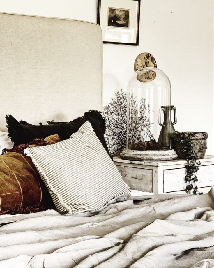 A cozy and rustic bedroom corner featuring an unmade bed with rumpled gray linens and an assortment of pillows in earthy tones. A wooden bedside table displays a cloche containing dried branches, flanked by a rustic pot and a glass jar, under an off-white lampshade. A small painting hangs above, contributing to the room's vintage charm.