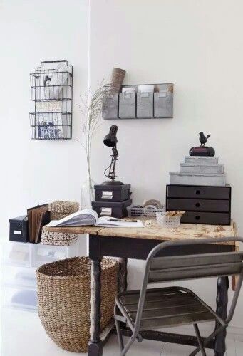 A well-organized home office corner with rustic wooden desk, vintage-style metal chair, and decorative storage solutions including a woven basket, stack of black drawers, and wall-mounted wire magazine rack.