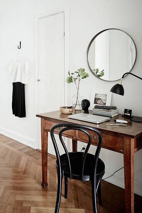An elegantly simple small living space with herringbone wood flooring. A classic wooden desk with a marble top is adorned with a small potted plant, a stack of books, and a laptop. A black bentwood chair is tucked under the desk. A striking round mirror hangs above, reflecting a white door with a stylish black and white outfit hanging on a hook beside it. The minimalist aesthetic is enhanced by the natural light and neutral tones of the room.